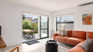 A modern living room with a sofa, a coffee table, and a black trash bin. There is a sliding glass door leading to an outdoor area with a table and chairs.
