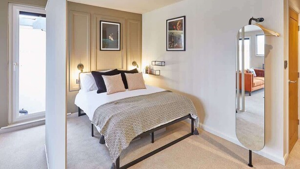 Un dormitorio con cama, mesitas de noche, un espejo y una puerta que da a otra habitación.