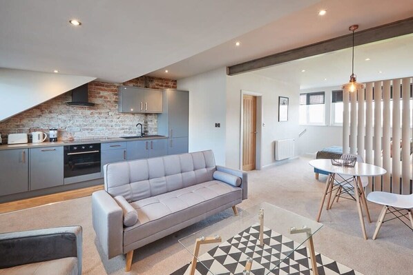 A modern kitchen with grey cabinets, a brick wall, and a grey sofa in a living area with a geometric rug.