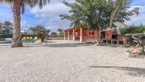A building with a red exterior, a covered outdoor area, and a pool with yellow chairs. - 016 - Finca La Dama Valverde - comfortHOLIDAYS (Valverde)