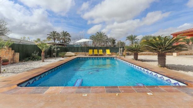 A rectangular swimming pool with a tiled edge, surrounded by a stone border and a tiled deck. There are three yellow lounge chairs aligned along the pool's edge. - 016 - Finca La Dama Valverde - comfortHOLIDAYS (Valverde)