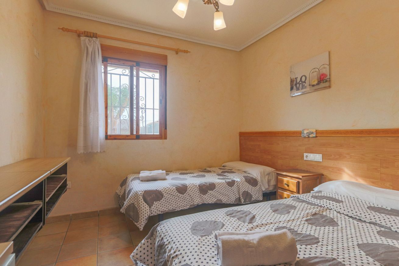 Slaapkamer met twee bedden, een houten hoofdbord, een nachtkastje, een raam met metalen tralies en een wandlamp.
