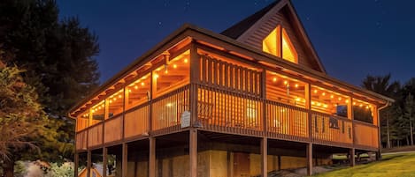 A two-story wooden cabin with a balcony, surrounded by a grassy area and trees.