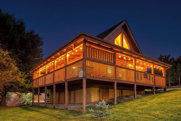 A two-story wooden cabin with a balcony, surrounded by a grassy area and trees.