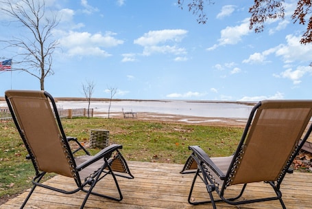 Two lounge chairs on a wooden deck overlooking a lake with a distant shoreline.