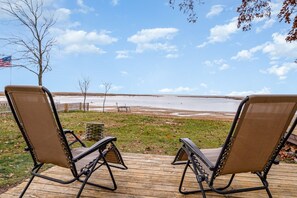 Dos sillones de relax en una terraza de madera con vistas a un lago y una orilla distante.