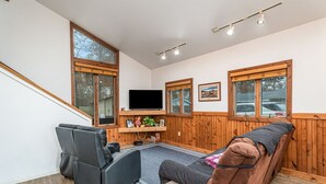 A cozy living room with a sofa, a chair, a flat-screen TV, wooden walls, and large windows. - Cozy Home on Lake Huron I (Au Gres)