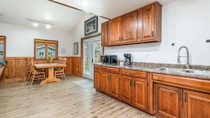A kitchen with wooden cabinets, a granite countertop, and a dining area with a wooden table and chairs. - Cozy Home on Lake Huron I (Au Gres)