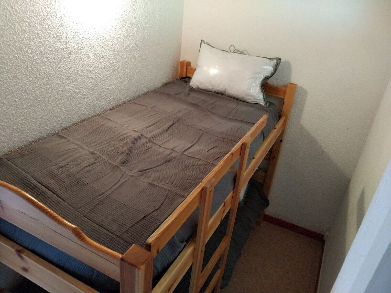 A small, single-bed room with a wooden frame, a gray mattress, and a white pillow.