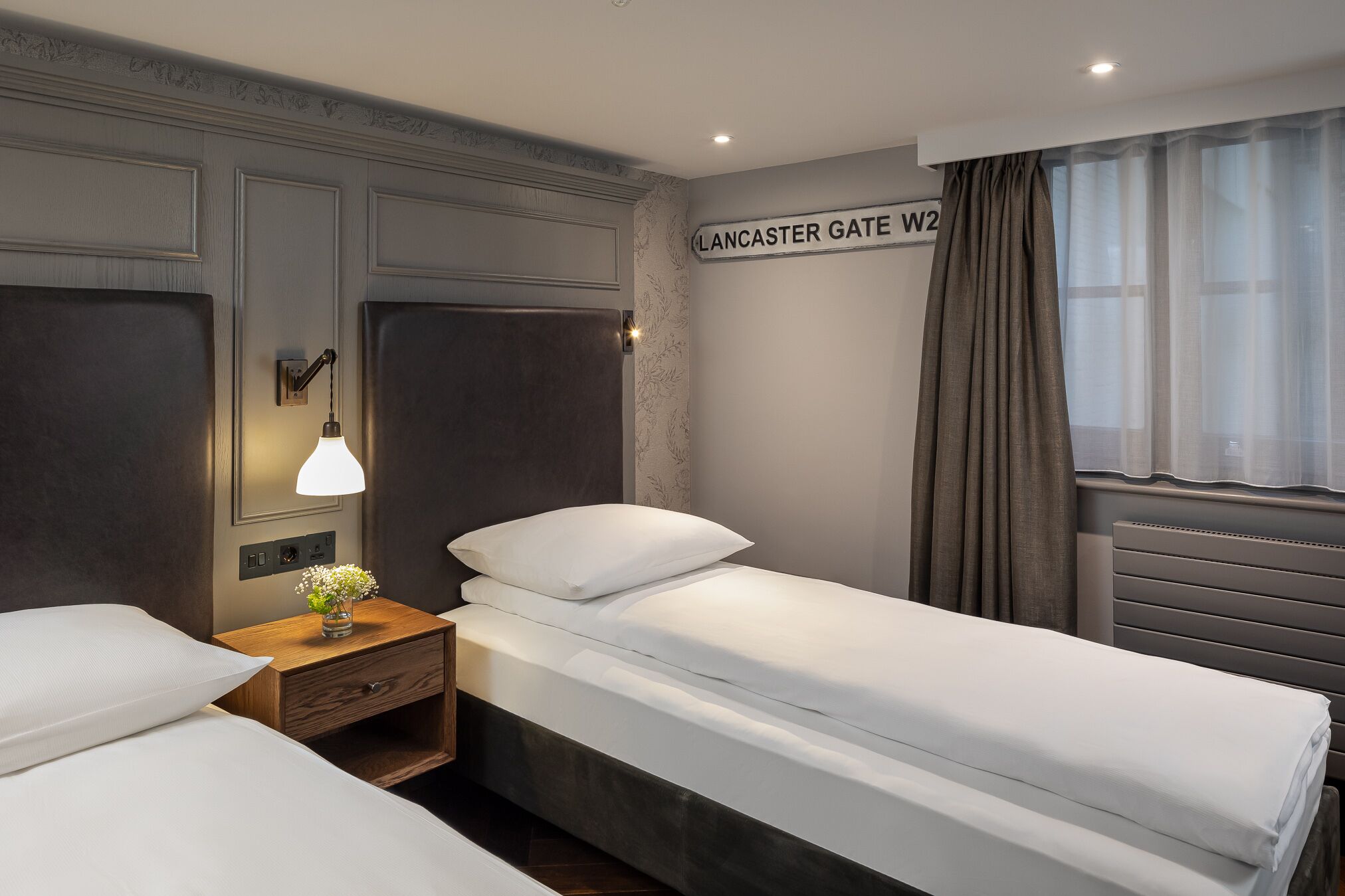 A hotel room with two single beds, a nightstand with a lamp, a wall sign reading "Lancaster Gate W2", and a window with curtains.
