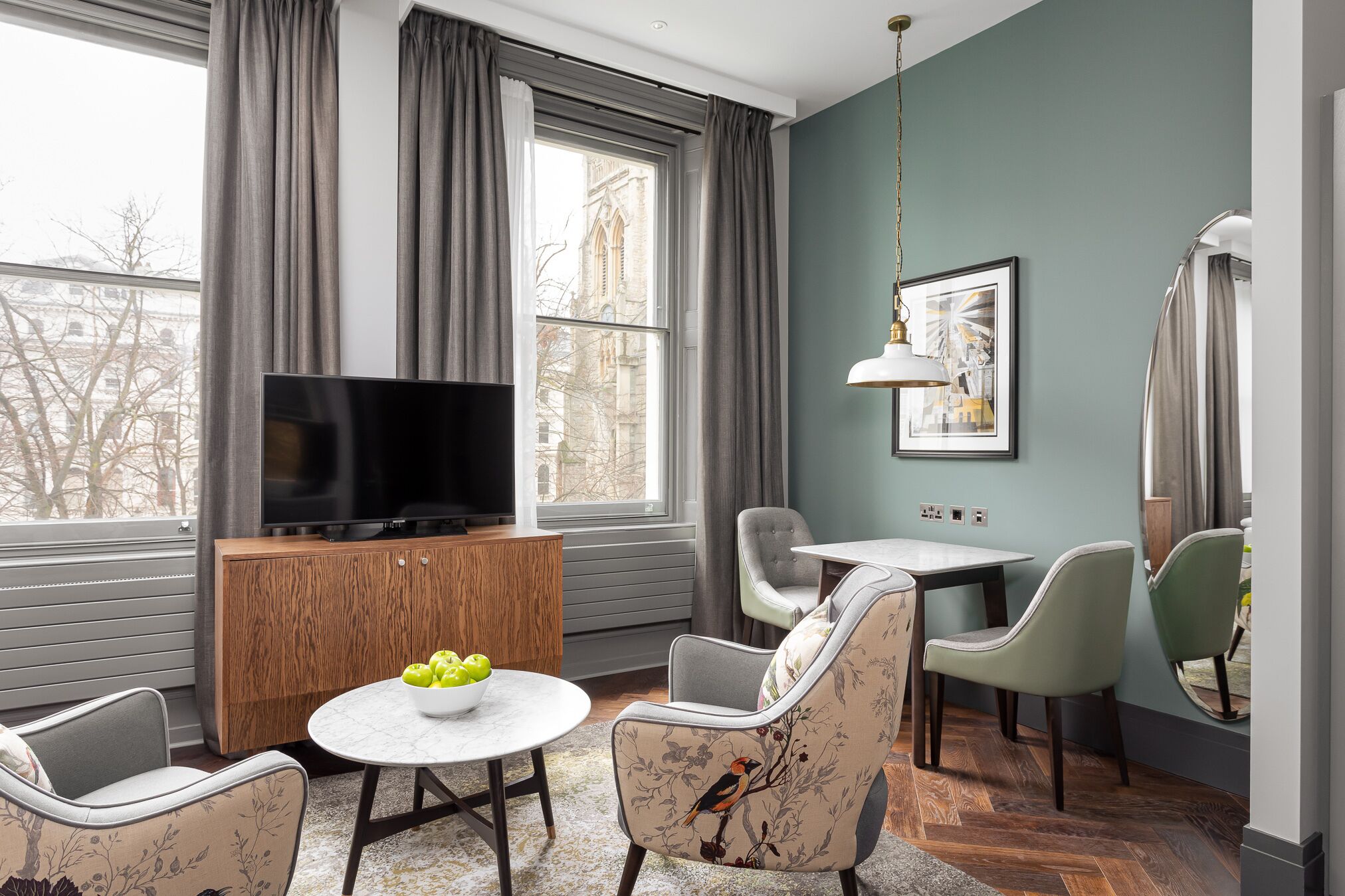 A modern living room with a flat-screen TV, wooden cabinet, round dining table, and two upholstered chairs.