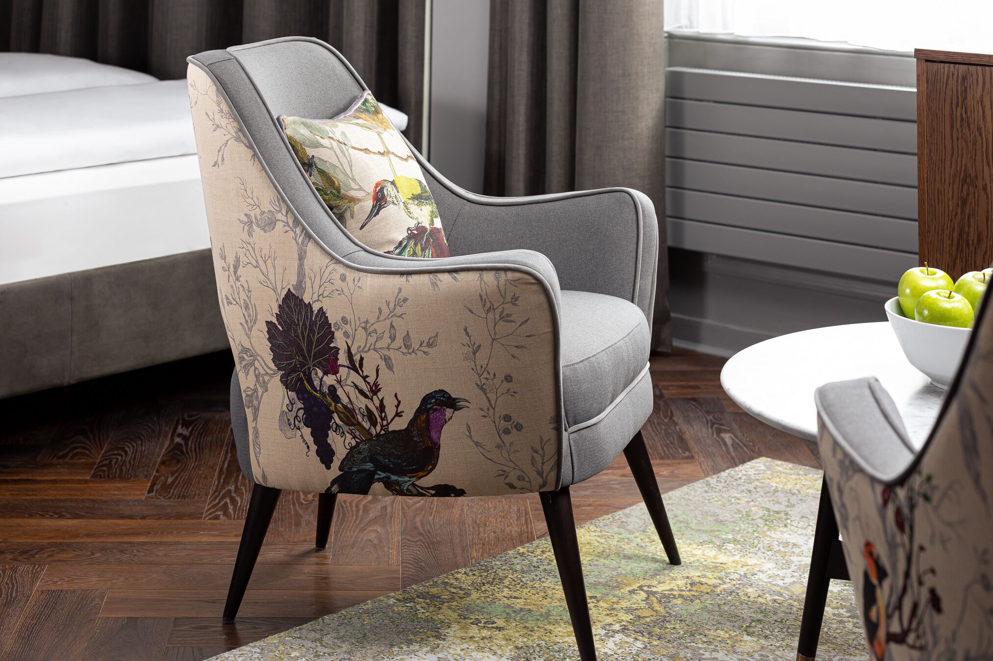 A modern living room with a grey upholstered armchair featuring a bird print, a white round table with a bowl of green apples, and a grey sofa in the background.