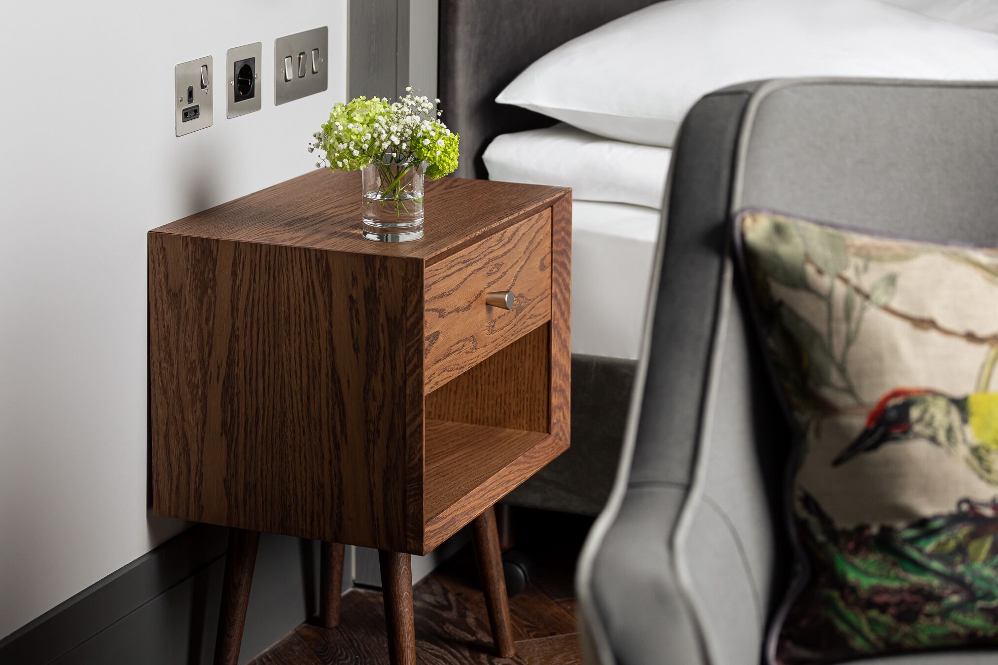 A small wooden side table with a glass vase containing white flowers, positioned next to a bed with a white pillow and a cushion with a bird design.