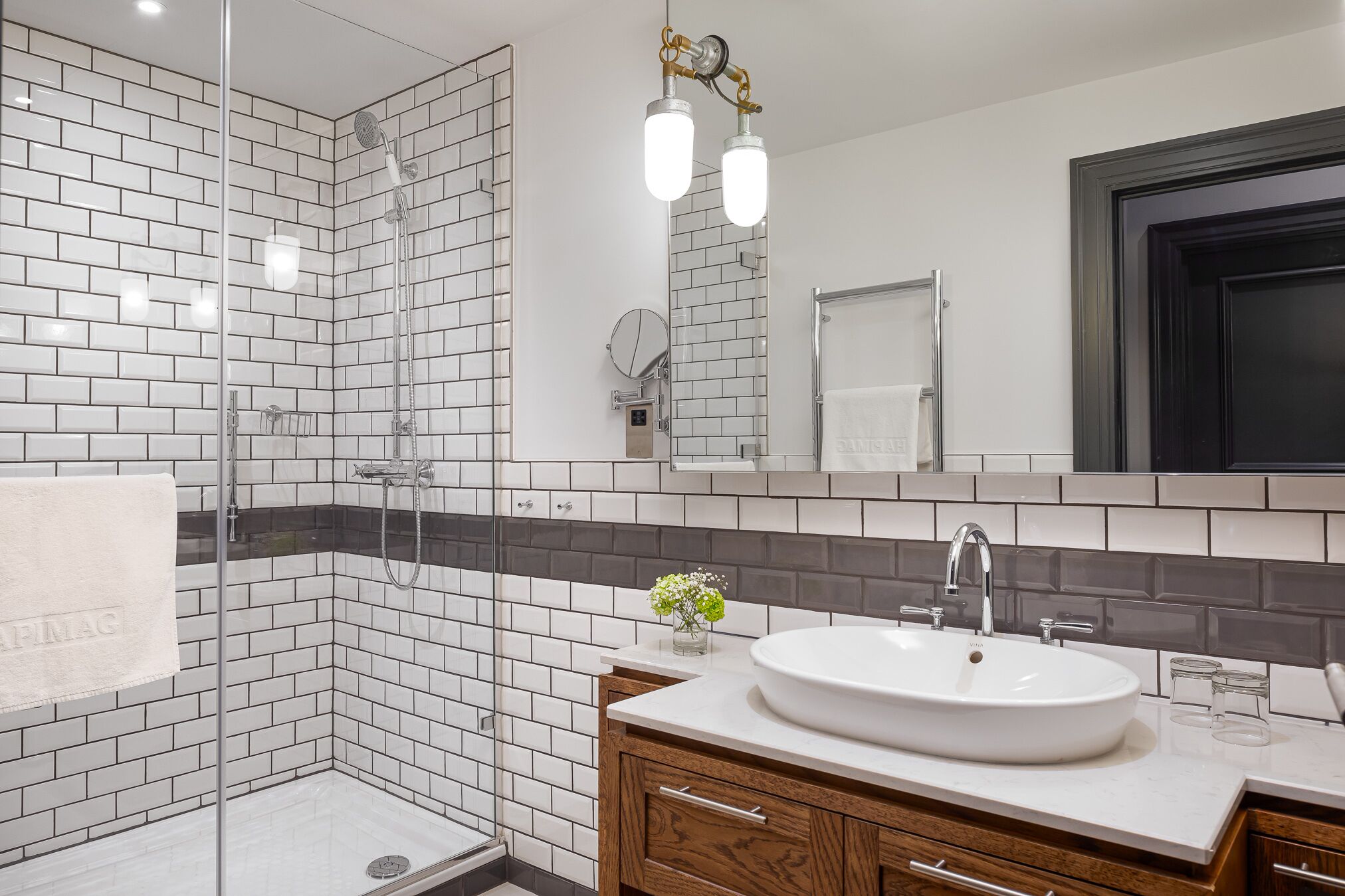 A modern bathroom with a glass-enclosed shower, white and gray tiled walls, a large mirror, and a white sink with wooden cabinets.