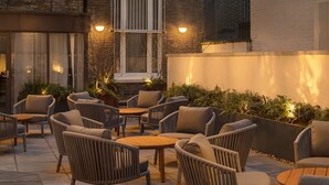 An outdoor seating area with wicker chairs and round tables, surrounded by plants and a stone wall. - Hapimag Apartments London (London)