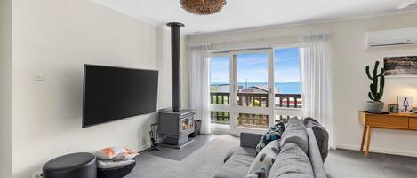 Ett vardagsrum med en stor platt-TV, en öppen spis, en soffa och utsikt över havet genom fönstren.