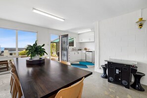 Eine moderne Küche mit weißen Schränken, ein Essbereich mit einem dunklen Holztisch und Stühlen sowie ein Blick aufs Meer durch große Fenster.