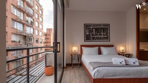 A hotel room with a bed, two bedside tables, a window with a view of other buildings, and a potted plant. - w* | Artistic Loft in Parque Virrey (Bogotá)