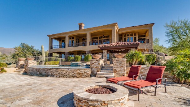A large, multi-story house with a patio area featuring a fire pit and red lounge chairs.