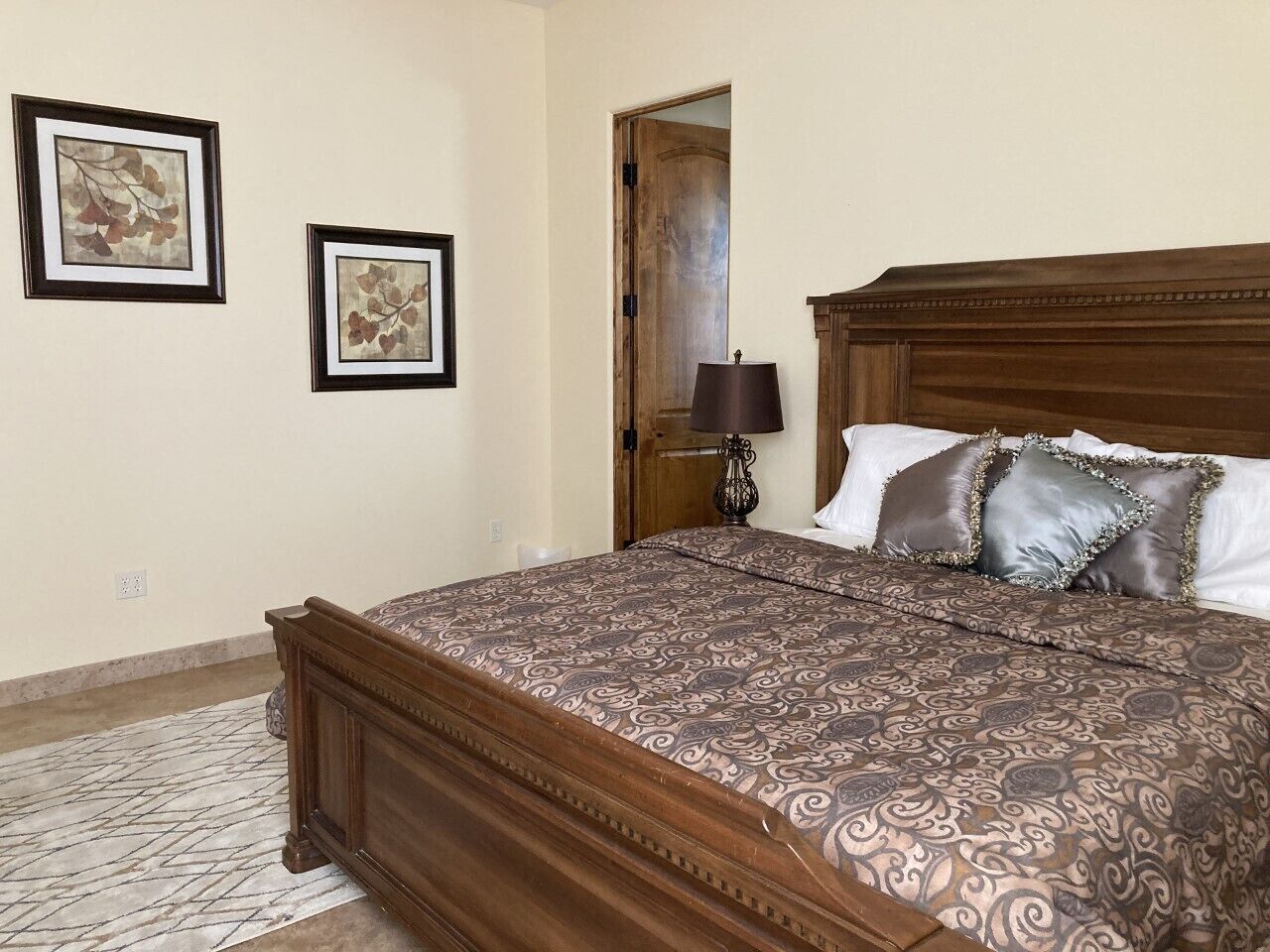 A bedroom with a wooden bed, patterned bedding, two framed artworks on the wall, and a nightstand with a lamp.