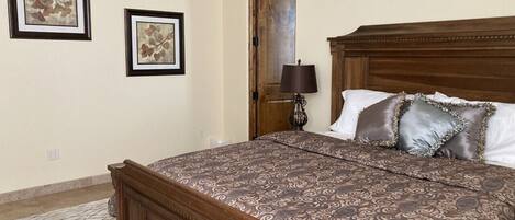A bedroom with a wooden bed, patterned bedding, two framed artworks on the wall, and a nightstand with a lamp.
