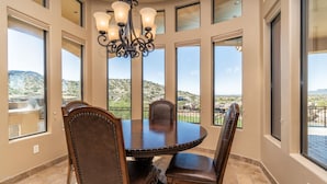 A dining area with a round table and four chairs, large windows with a view of a hilly landscape, and a chandelier.