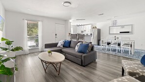 Ein modernes Wohnzimmer mit einem grauen Sofa, einem runden Couchtisch und einem Essbereich mit weißem Tisch und Stühlen.