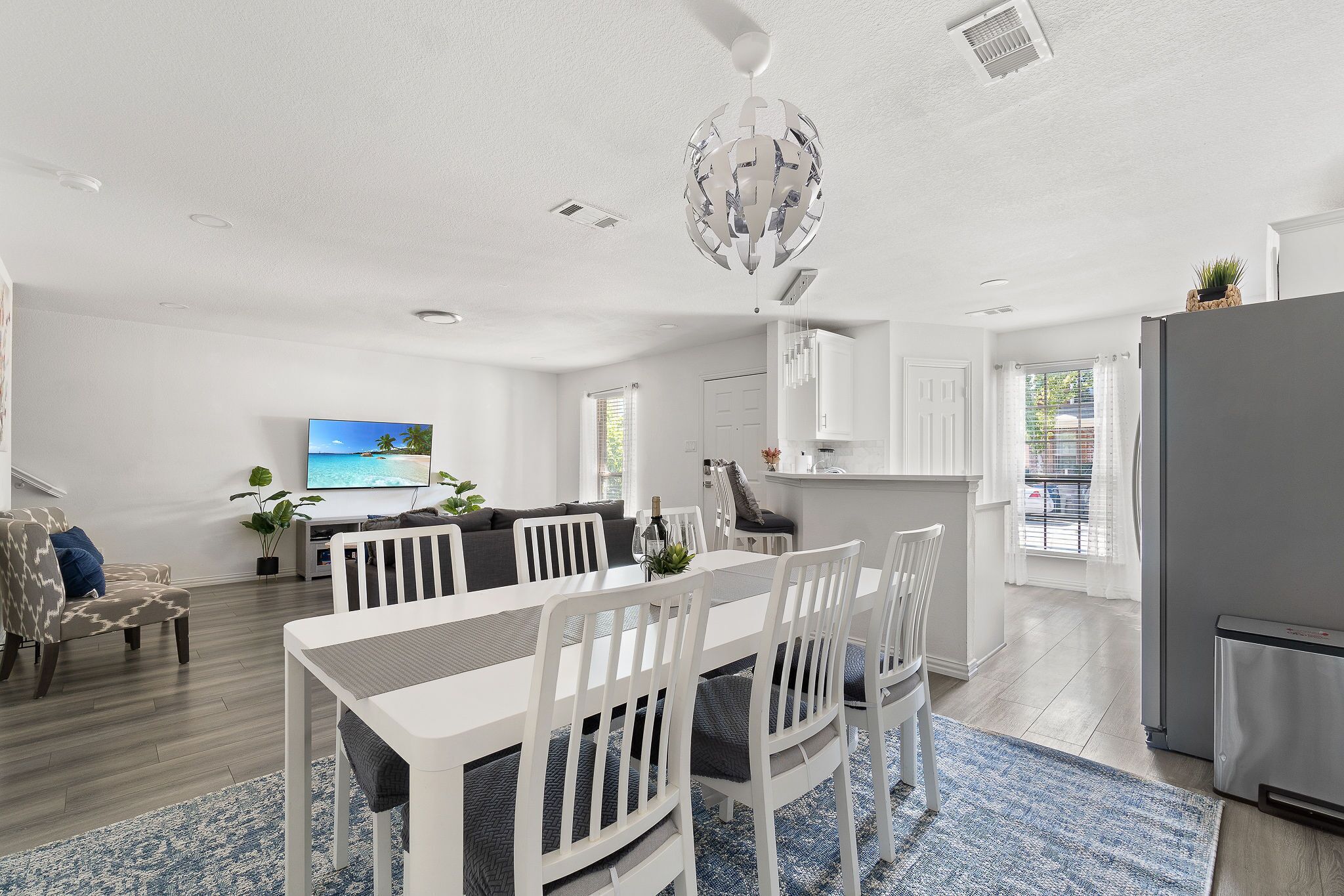 A modern living room with a dining area, a kitchen, and a TV displaying a scenic view.