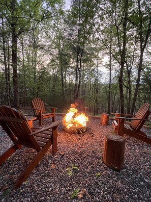 A forest with a fire pit surrounded by wooden chairs and a wooden stump.