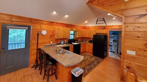 A wooden cabin kitchen with a central island, bar stools, a refrigerator, and a window overlooking a forest.