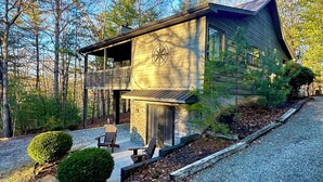 A two-story log cabin with a balcony, stone foundation, and a gravel driveway.