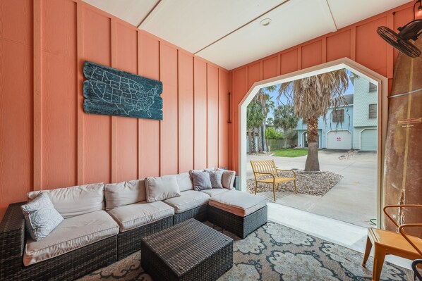 A cozy living area with a sectional sofa, two ottomans, and a wooden chair. A map artwork is on the wall, and there is a view of a tree and a house outside. - A Pet Friendly Tropical Oasis! (Port Aransas)