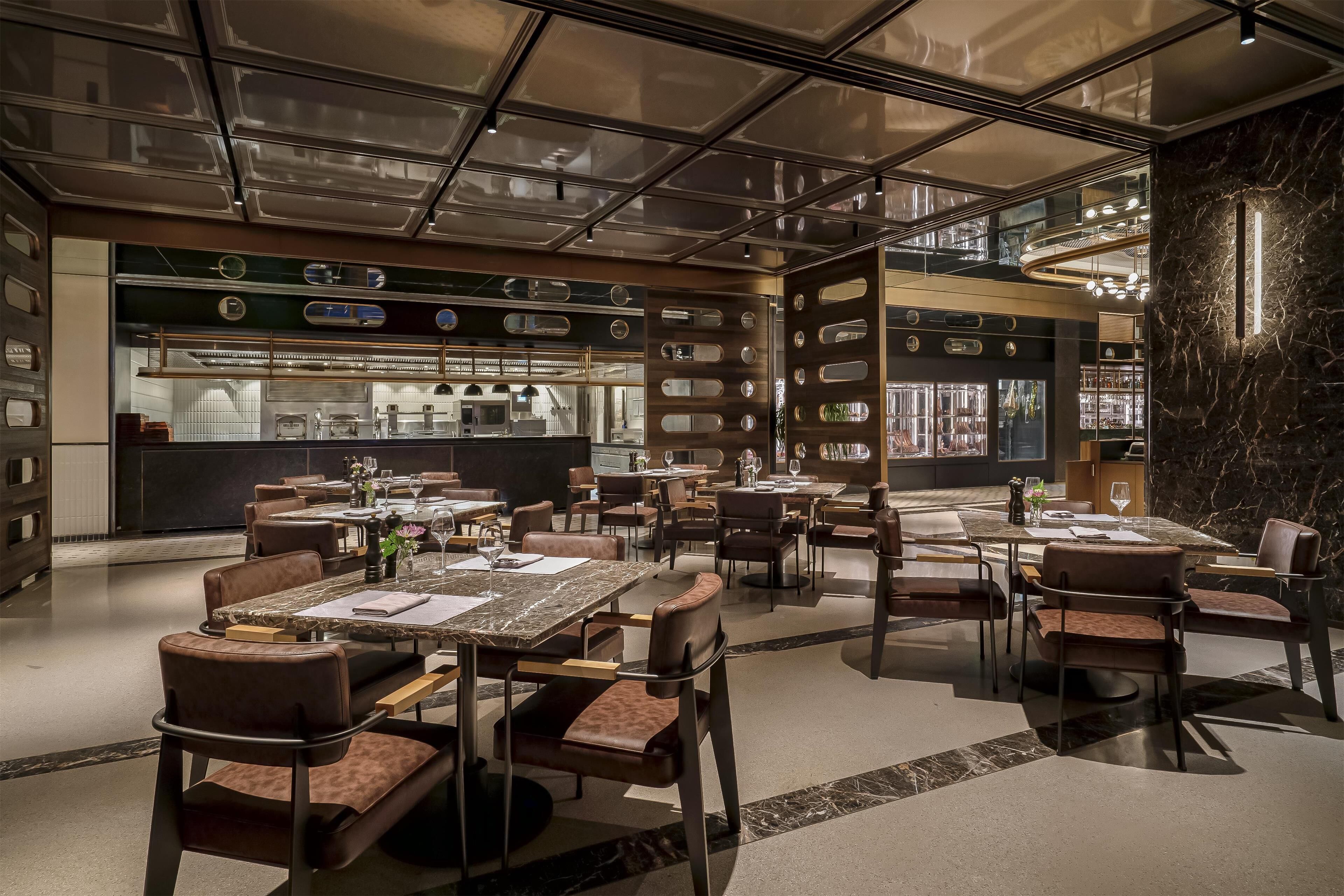 A modern restaurant with multiple dining tables and booths, a kitchen area in the background, and a stone wall with a light fixture.