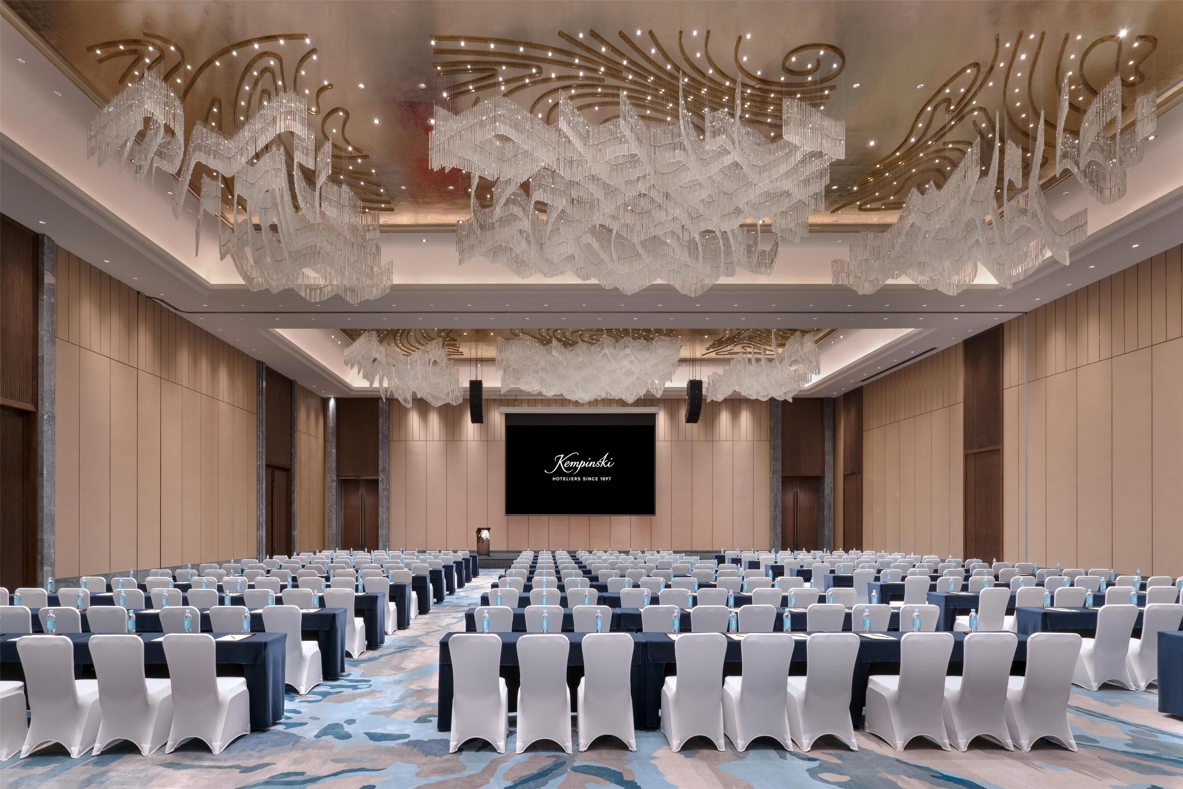 A conference room with rows of tables and chairs, a large screen, and a chandelier.
