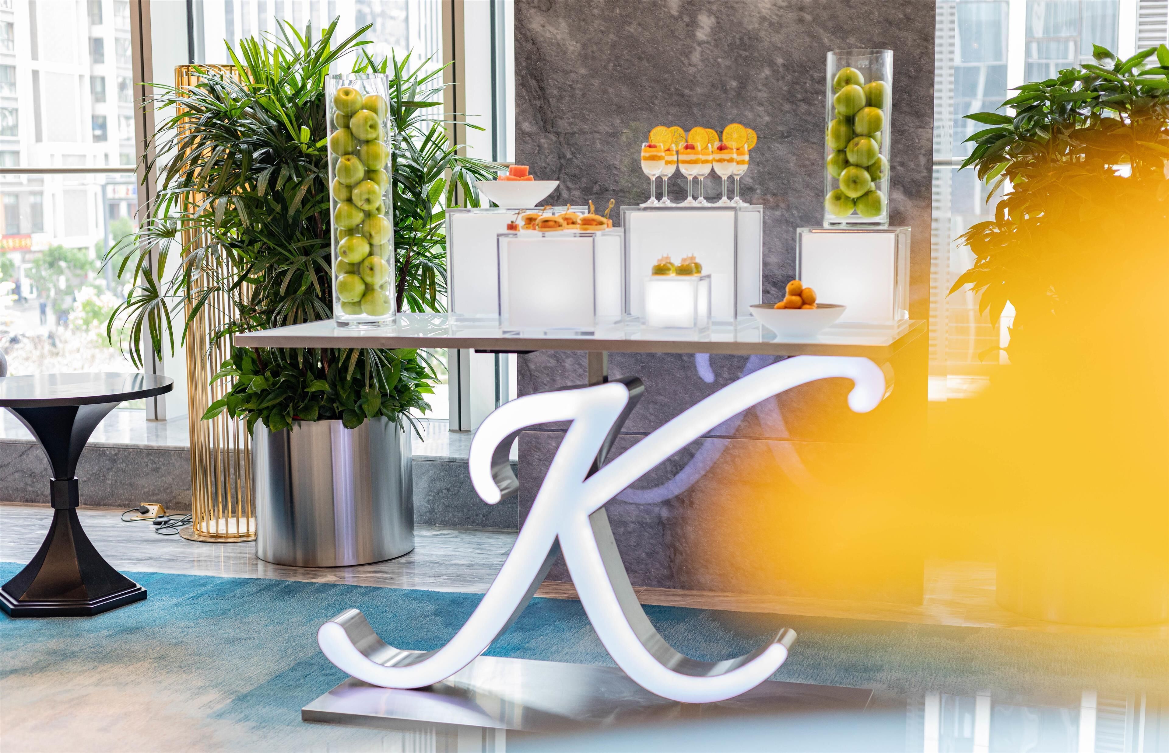 A modern hotel reception area with a glass display case, potted plants, and a large decorative letter 'K'.