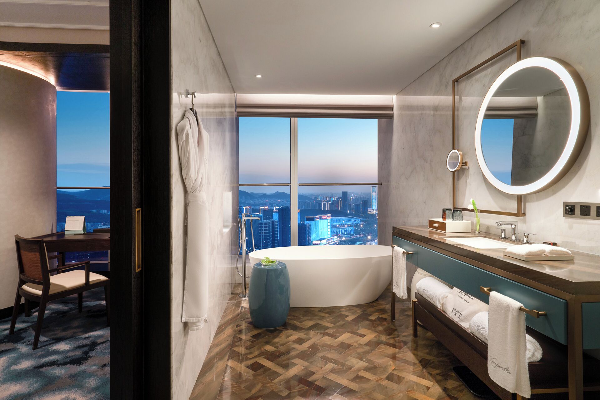 A modern hotel bathroom with a large bathtub, a vanity with a mirror, and a view of a cityscape through large windows.