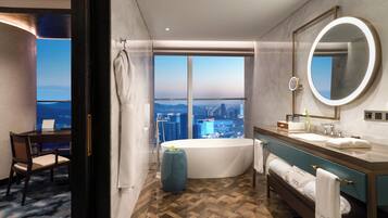 A modern hotel bathroom with a large bathtub, a vanity with a mirror, and a view of a cityscape through large windows.