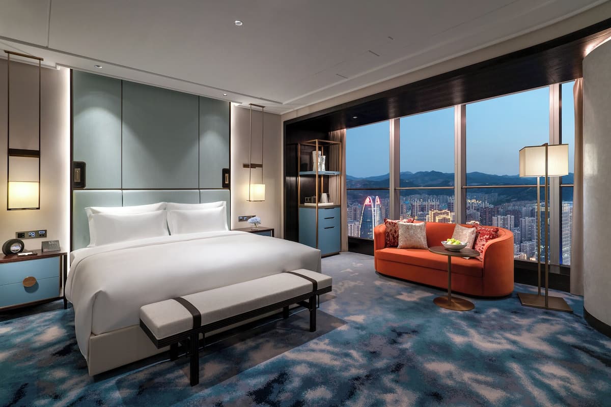 Une chambre d’hôtel moderne dotée d’un grand lit, d’une banquette, d’un canapé et offrant une vue sur le paysage urbain grâce à de grandes fenêtres.
