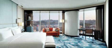 A hotel room with a large bed, a sofa, a desk, and a view of a cityscape through large windows.