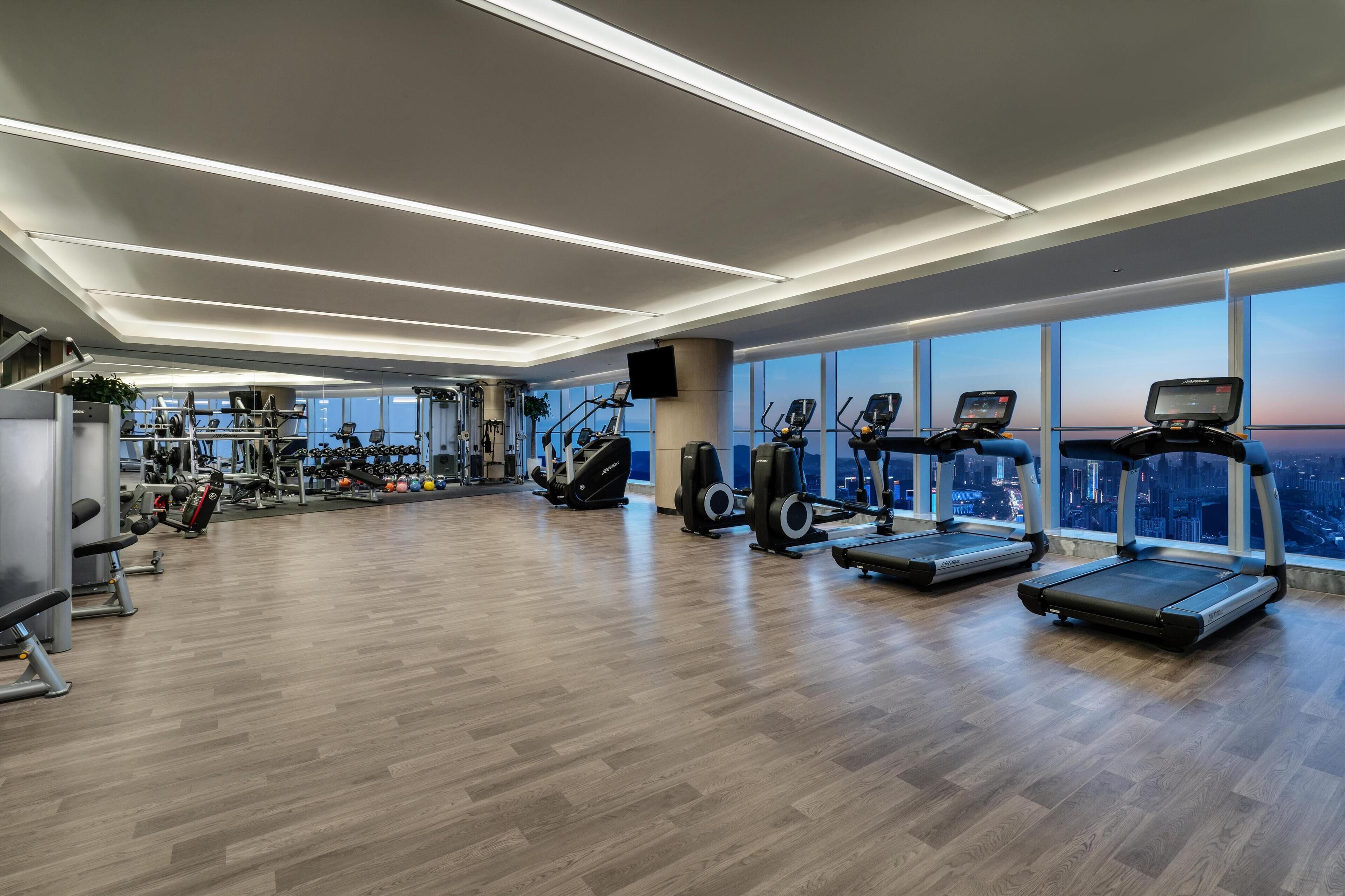 A modern gym with treadmills, weight machines, and a large window offering a city view.