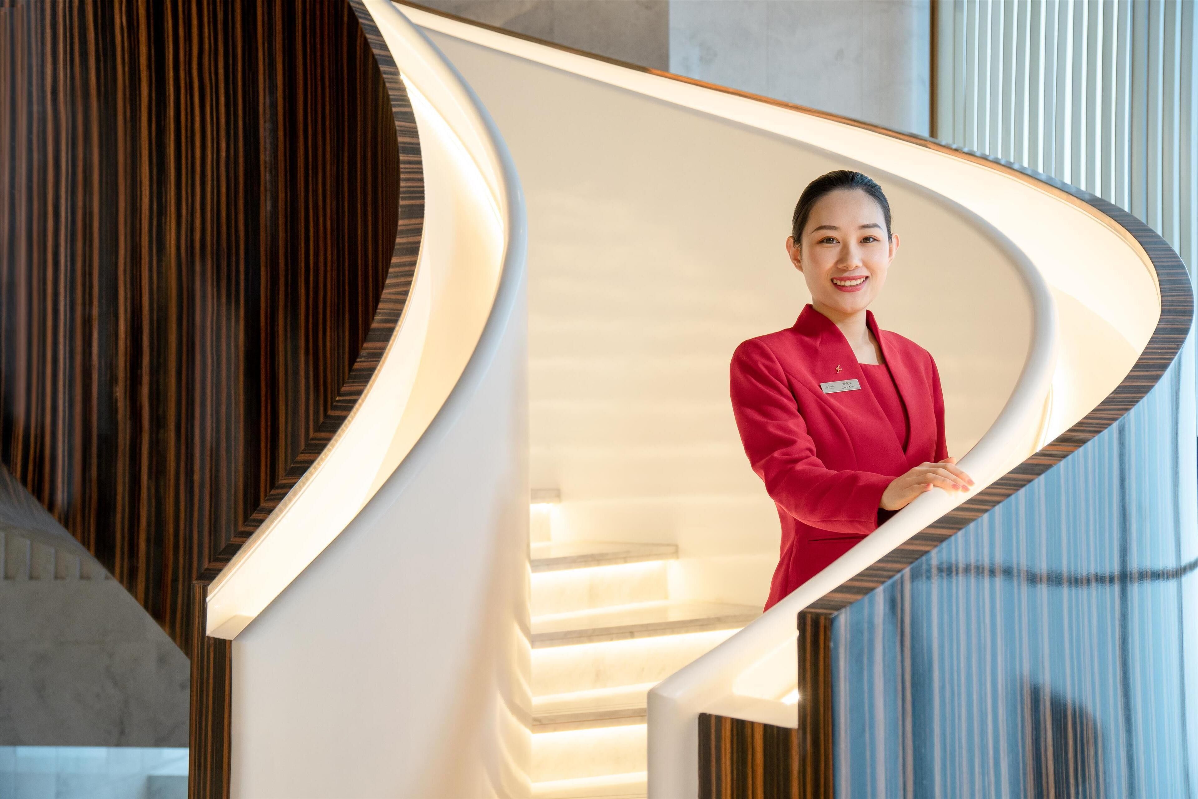 A hotel reception area with a modern design, featuring a curved reception desk and a staircase with wooden accents.