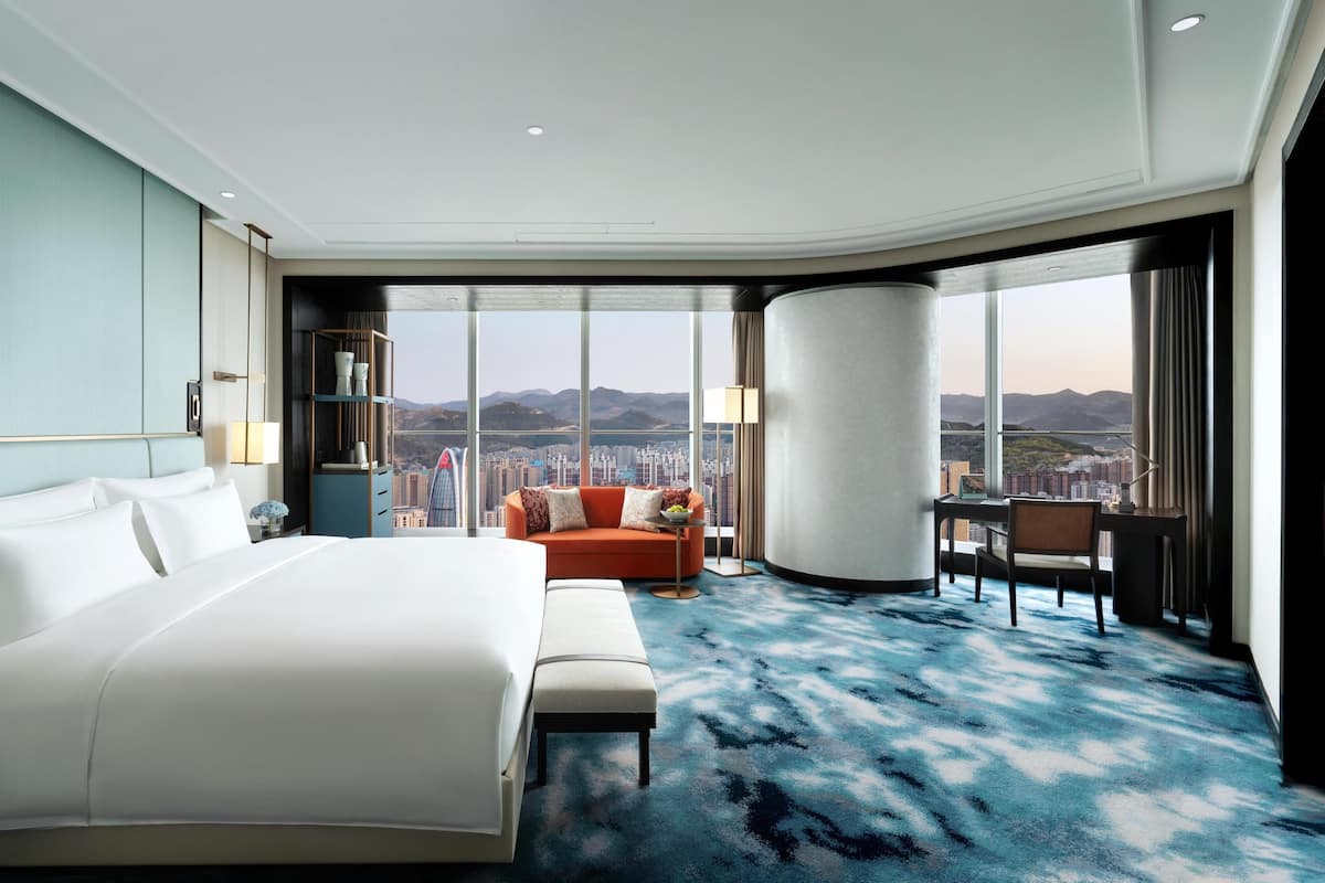Une chambre d’hôtel avec un grand lit, un canapé, un bureau et une vue sur le paysage urbain grâce à de grandes fenêtres.