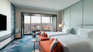 A hotel room with a large bed, a sofa, a small table, and a TV. The room has a large window with a city view.