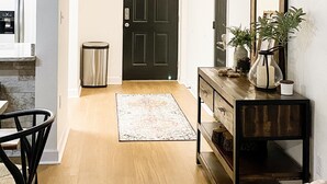 Un couloir avec une porte noire, une console en bois surmontée d’un miroir, une tête de cerf accrochée au mur et une poubelle.
