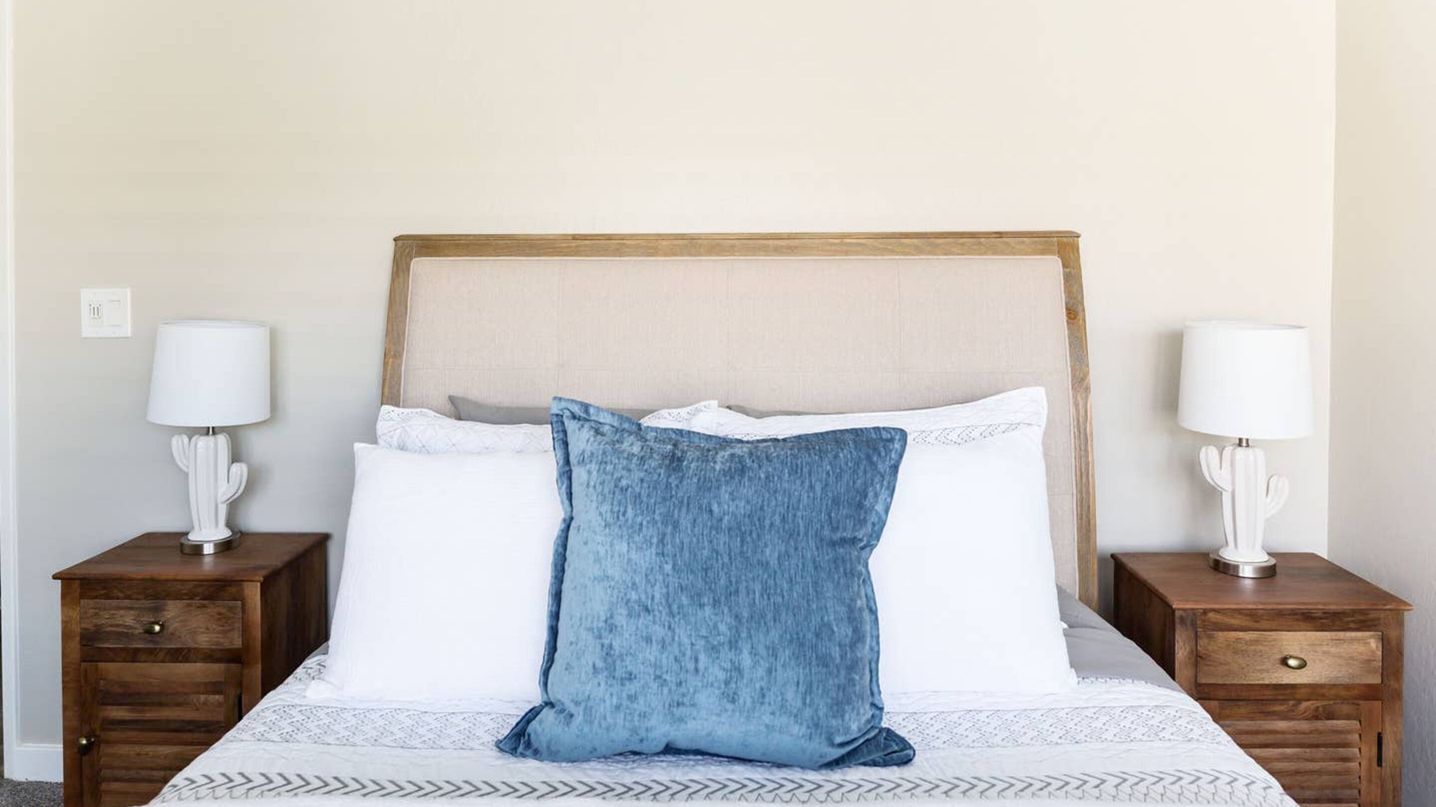 Una cama con cabecera de madera, ropa de cama blanca y almohada azul. Hay dos mesitas de noche con lámparas y un cajón.