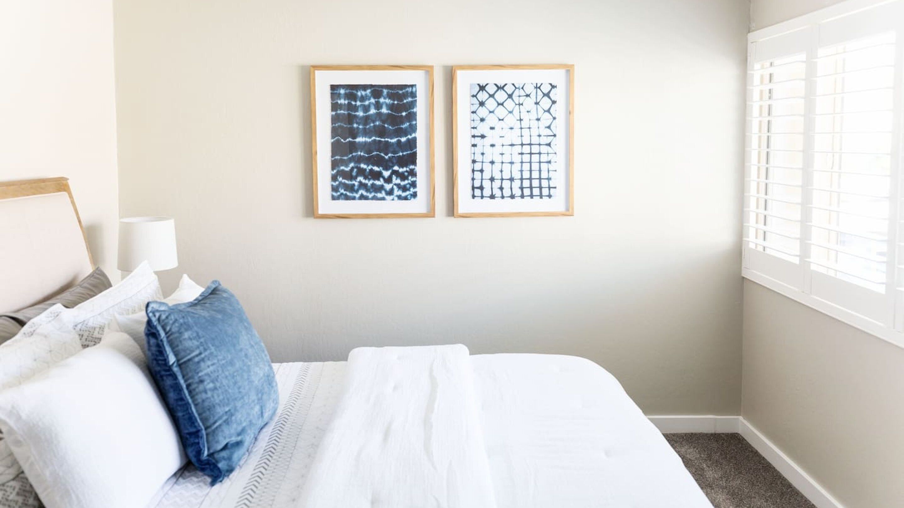 A neatly made bed with white linens and a blue pillow, two framed abstract artworks on the wall, and a window with blinds.