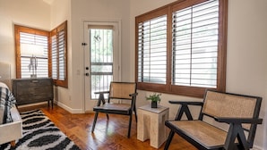 Ein Zimmer mit Holzboden, einer Tür mit Glasfenster, zwei Korbstühle, einem kleinen Tisch mit Pflanze und einem schwarz-weißen Teppich.