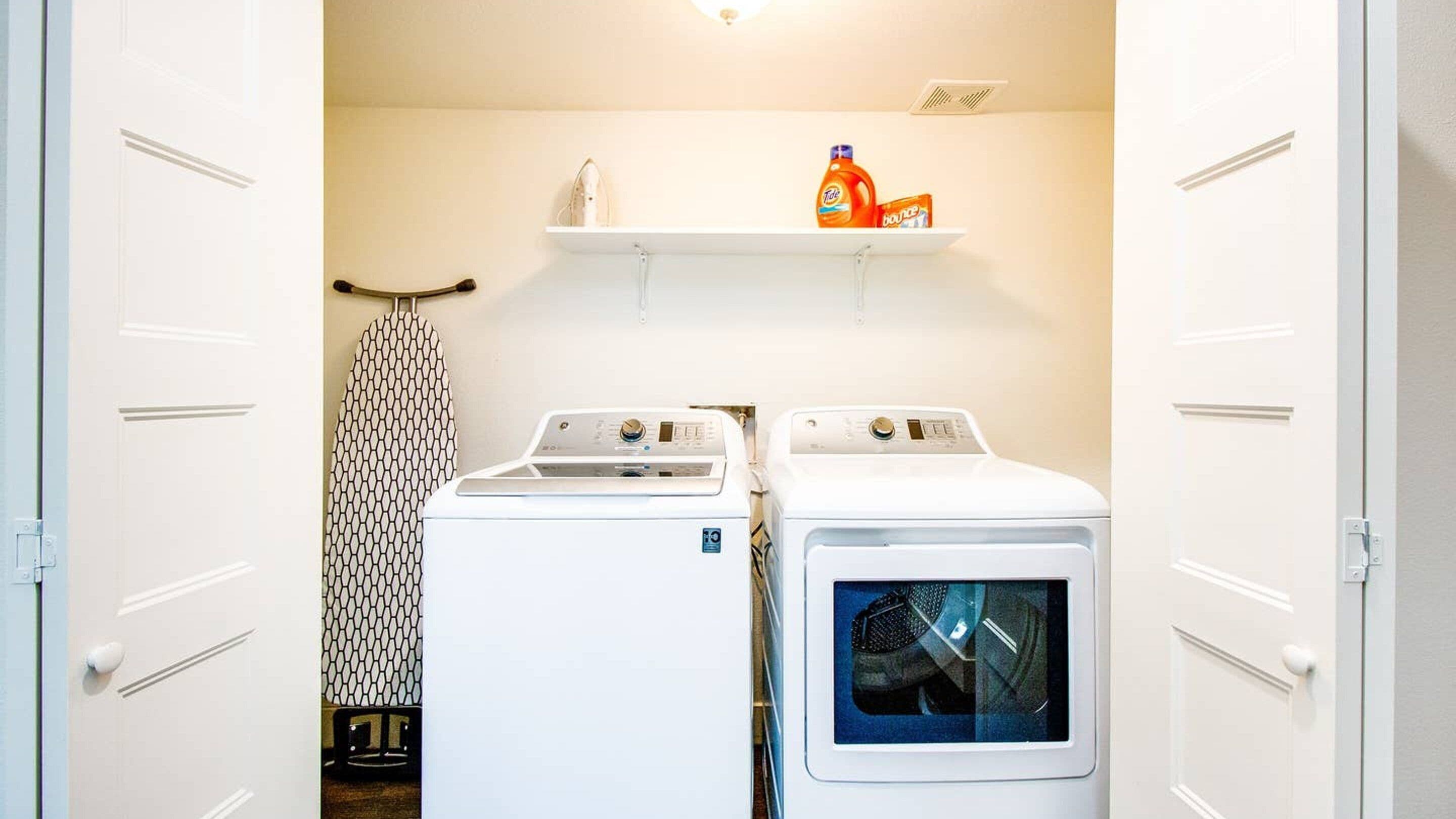A laundry room with two white washing machines, a shelf with detergent bottles, and a patterned ironing board.