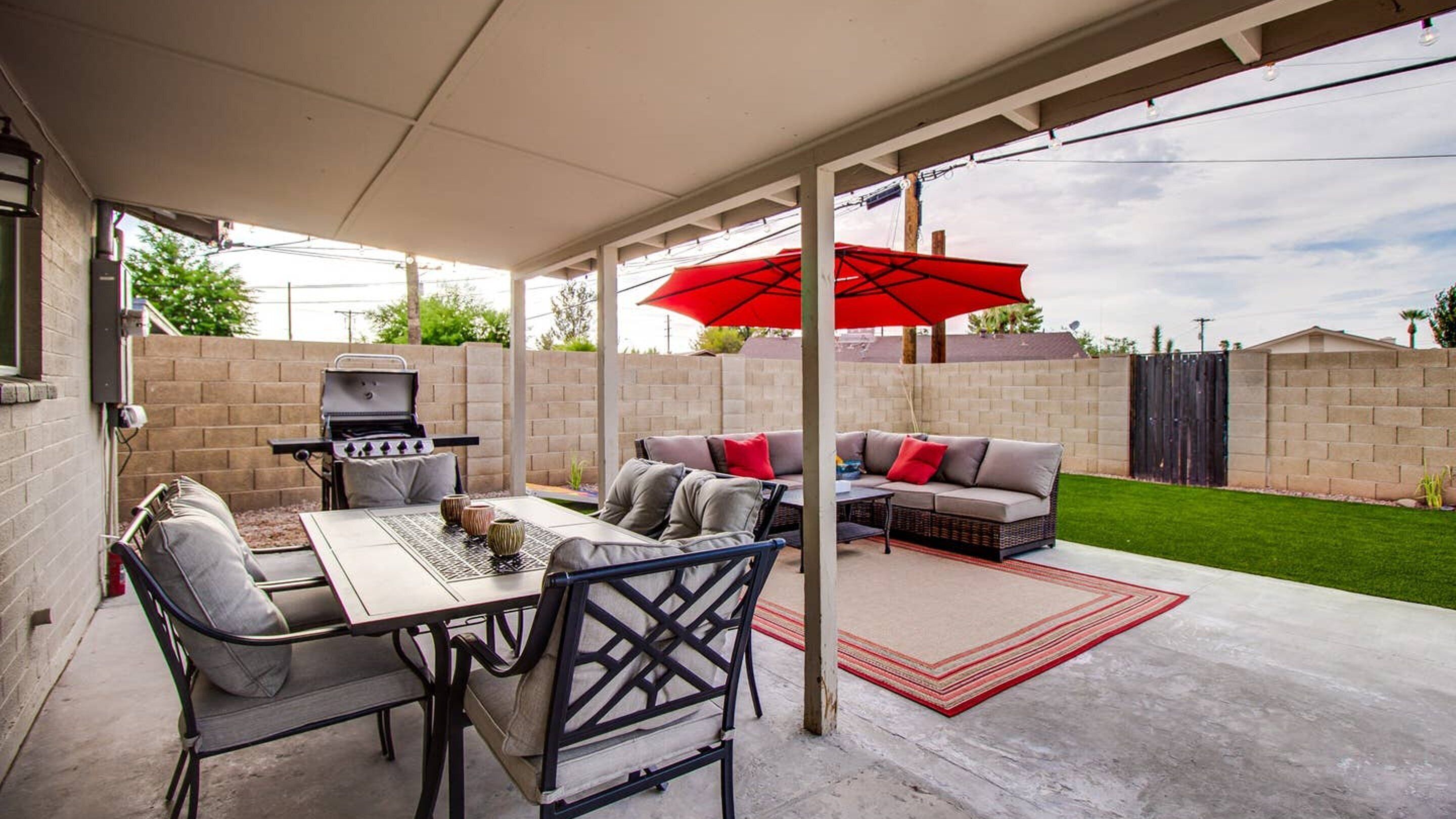 Une terrasse extérieure avec un barbecue, une table à manger, des chaises et un canapé placés sous un parasol rouge.
