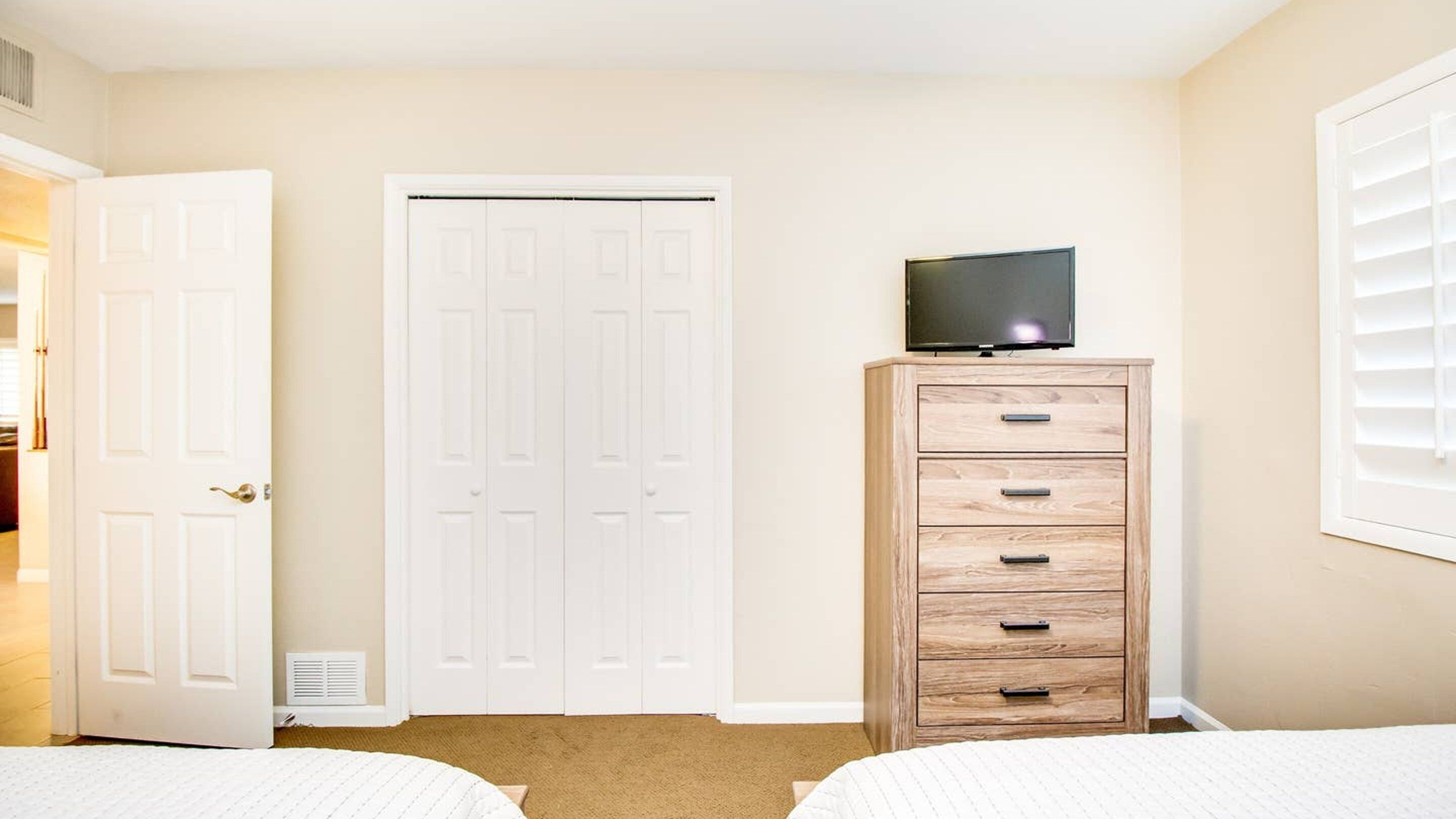 Un dormitorio con dos camas, un aparador de madera con un televisor encima, una puerta blanca y una ventana con persianas.